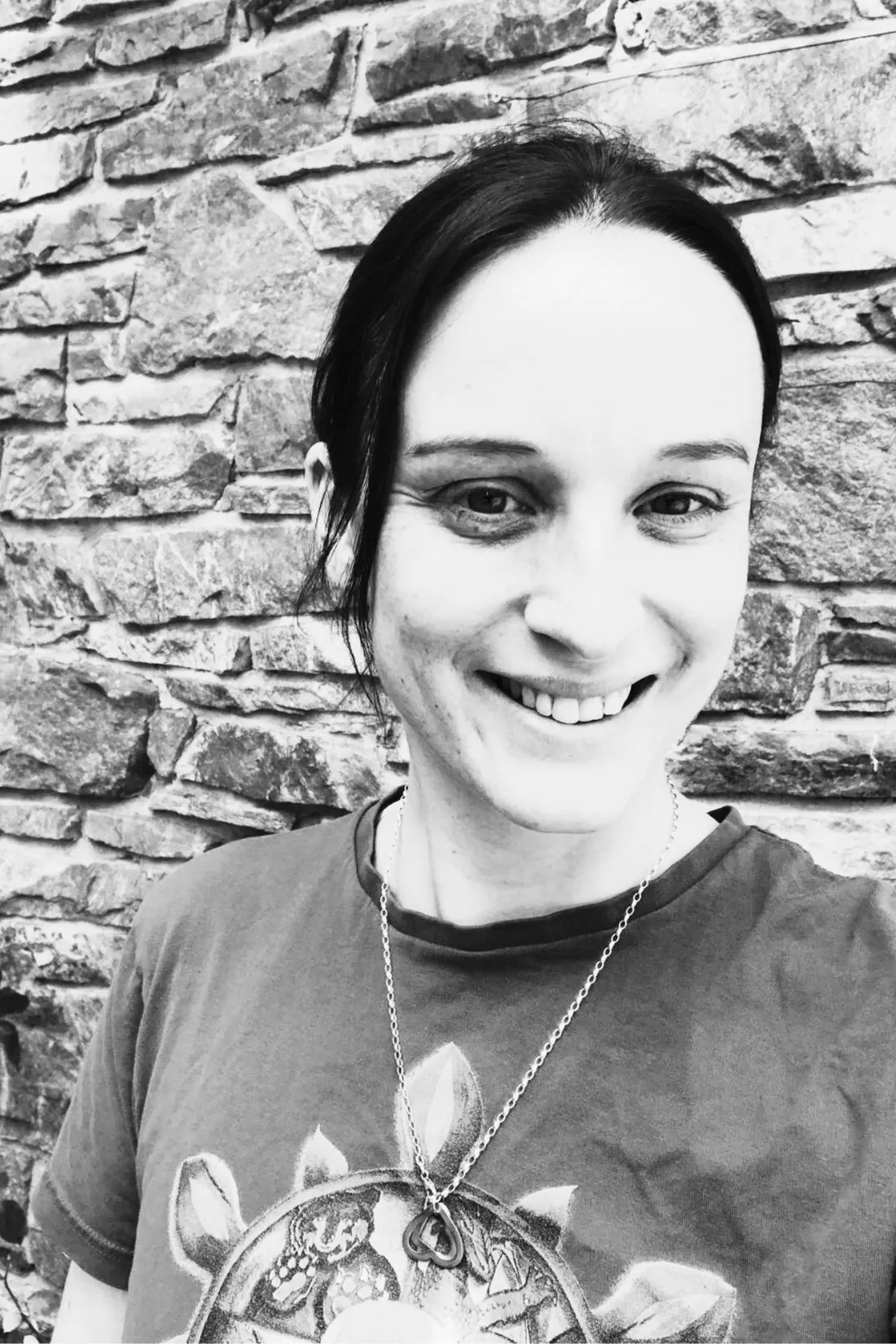 Schwarzweiß-Porträt: Sarah Karle lächelt vor einer Natursteinmauer in die Kamera, auf ihrem Shirt das Rad des Kung-Fu.