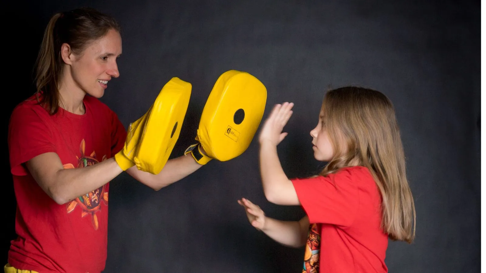 Ein Mädchen im roten WingTsun-Shirt holt mit beiden Händen aus und schlägt auf zwei gelbe Schlagpolster, die die lachende Trainerin ihr entgegenhält.